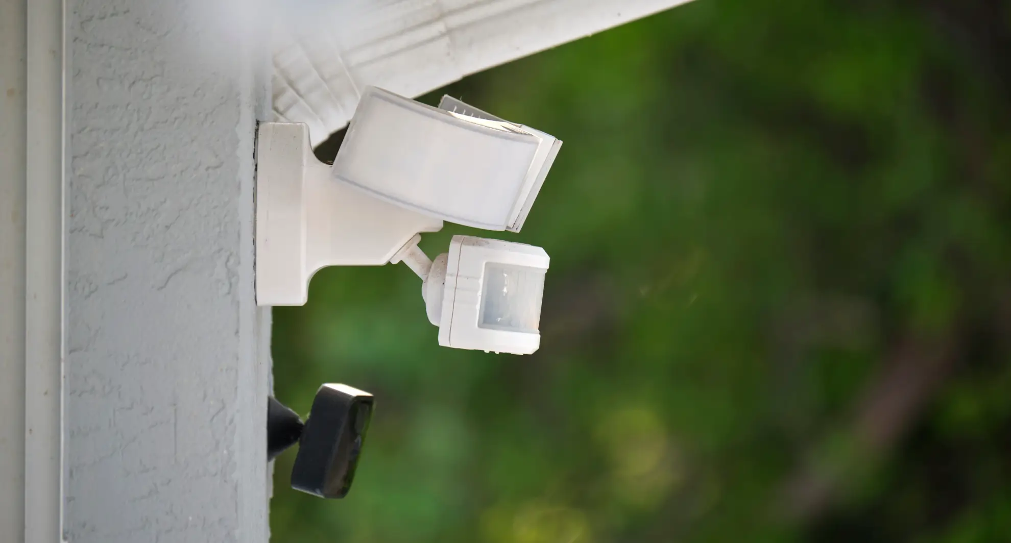 Close-up view of a white motion sensor security light mounted on the corner of a house exterior, with a blurred green foliage background, focusing on the shape and details of the fixture.