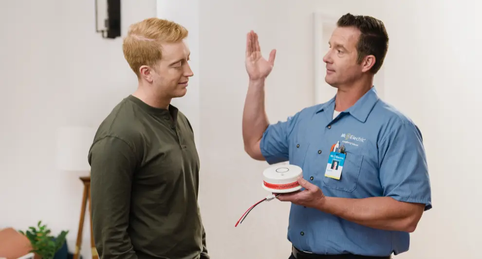An electrician in a blue uniform is explaining the features of a carbon monoxide detector to a homeowner in a modern living space.