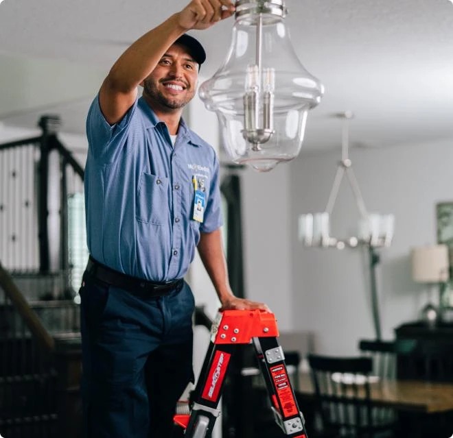 Smiling Mr. Electric electrician on a ladder adjusting a pendant light during a lighting services appointment