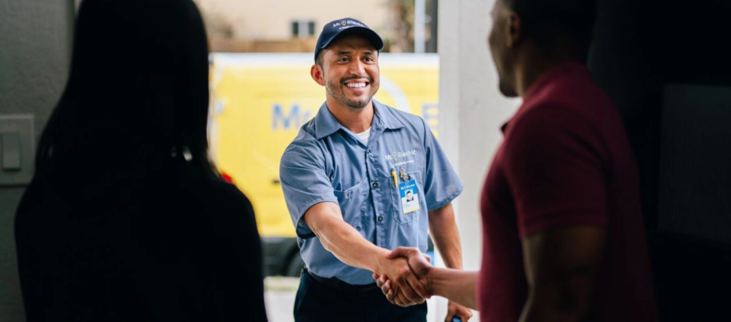 Smiling male Mr. Electric electrician shaking hands with customer silhouetted inside doorway.