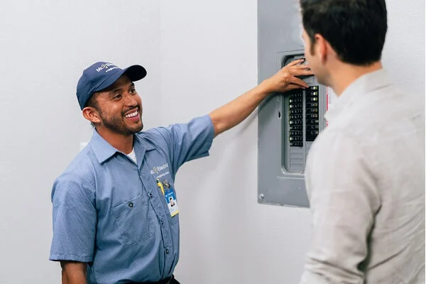 A smiling Mr. Electric electrician points at an open electrical panel while talking to a man with his back to the camera    